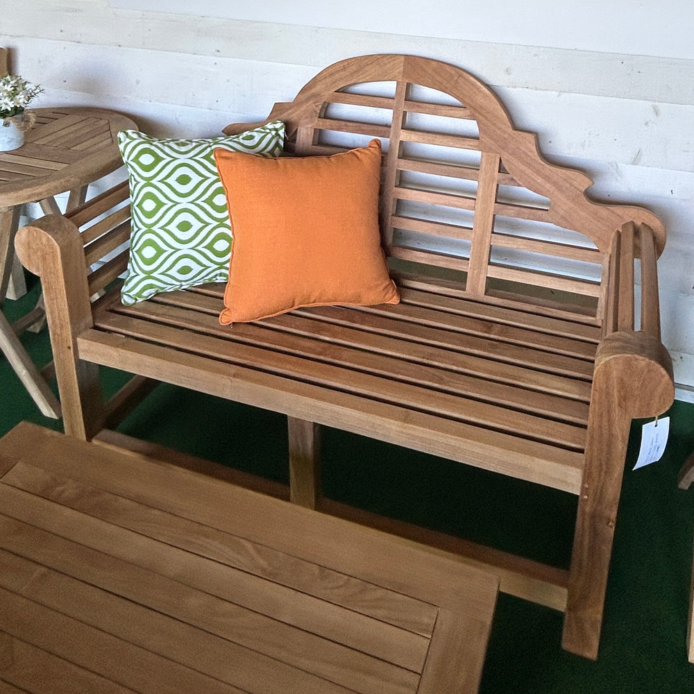 
                  
                    Teak bench with two pillows, one green and one orange, against a white wall, with teak tables to the side and in front.
                  
                