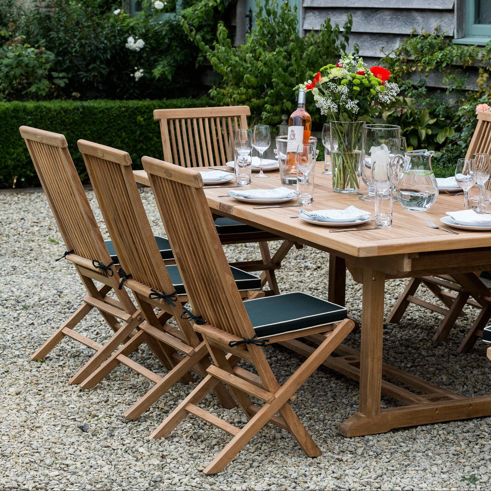 
                  
                    Outdoor dining setup with teak table and chairs in front of a rustic building.
                  
                