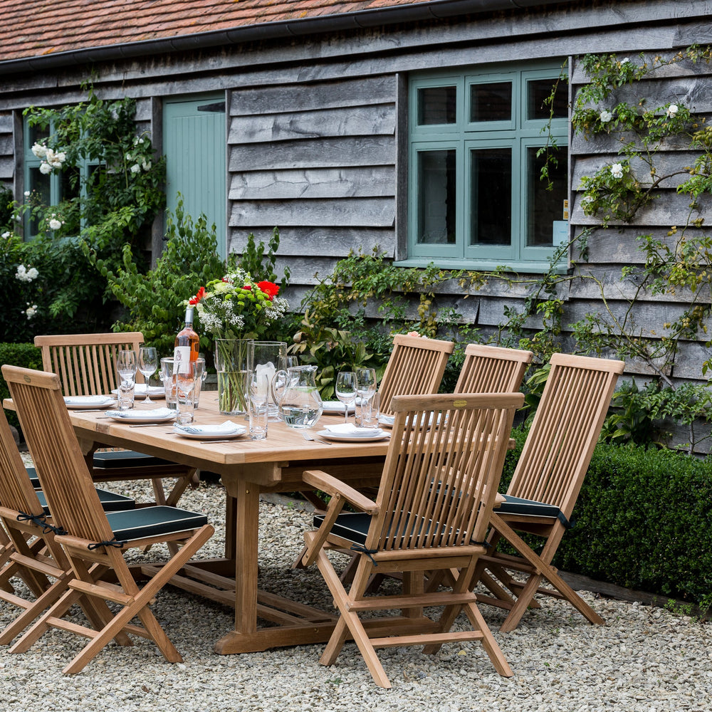
                  
                    Teak outdoor dining set with chairs on a patio in front of a rustic building.
                  
                