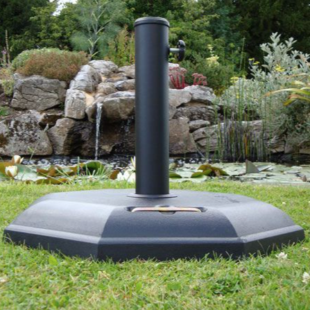 A black octagonal poly-concrete moulded parasol base with a steel tube and plastic reducer, set on grass with a waterfall and rocks in the background.