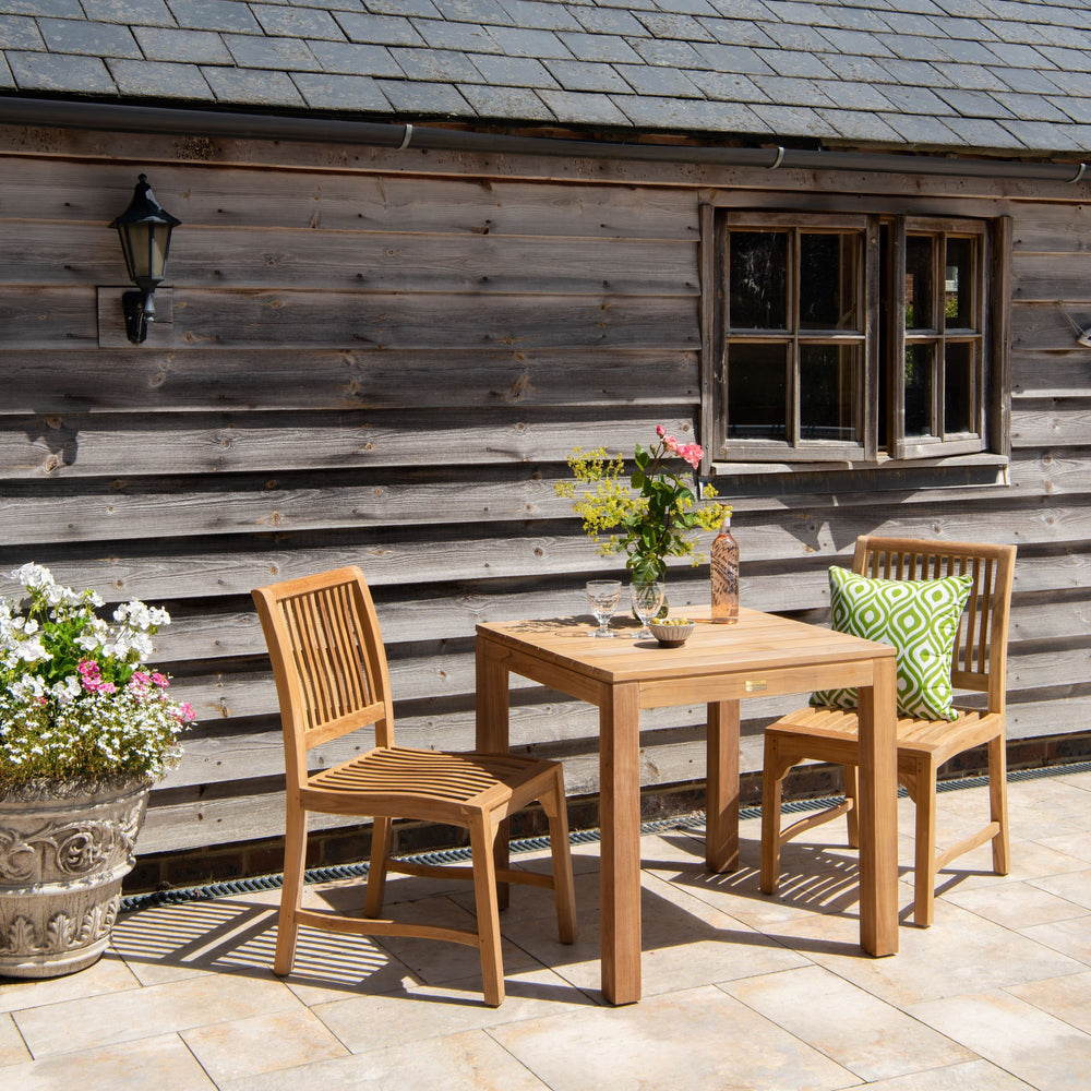 Teak outdoor table with two teak chairs and flowers in front of a rustic wooden building.