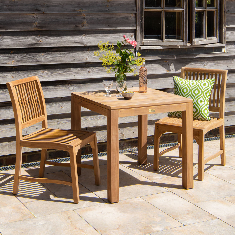 Teak outdoor table with two chairs, one with a green patterned cushion, on a patio, against a wooden wall.