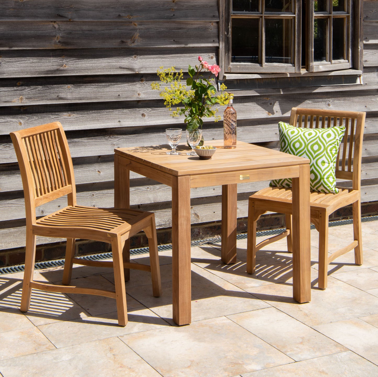 Teak outdoor table with two chairs, one with a green patterned cushion, on a patio, against a wooden wall.