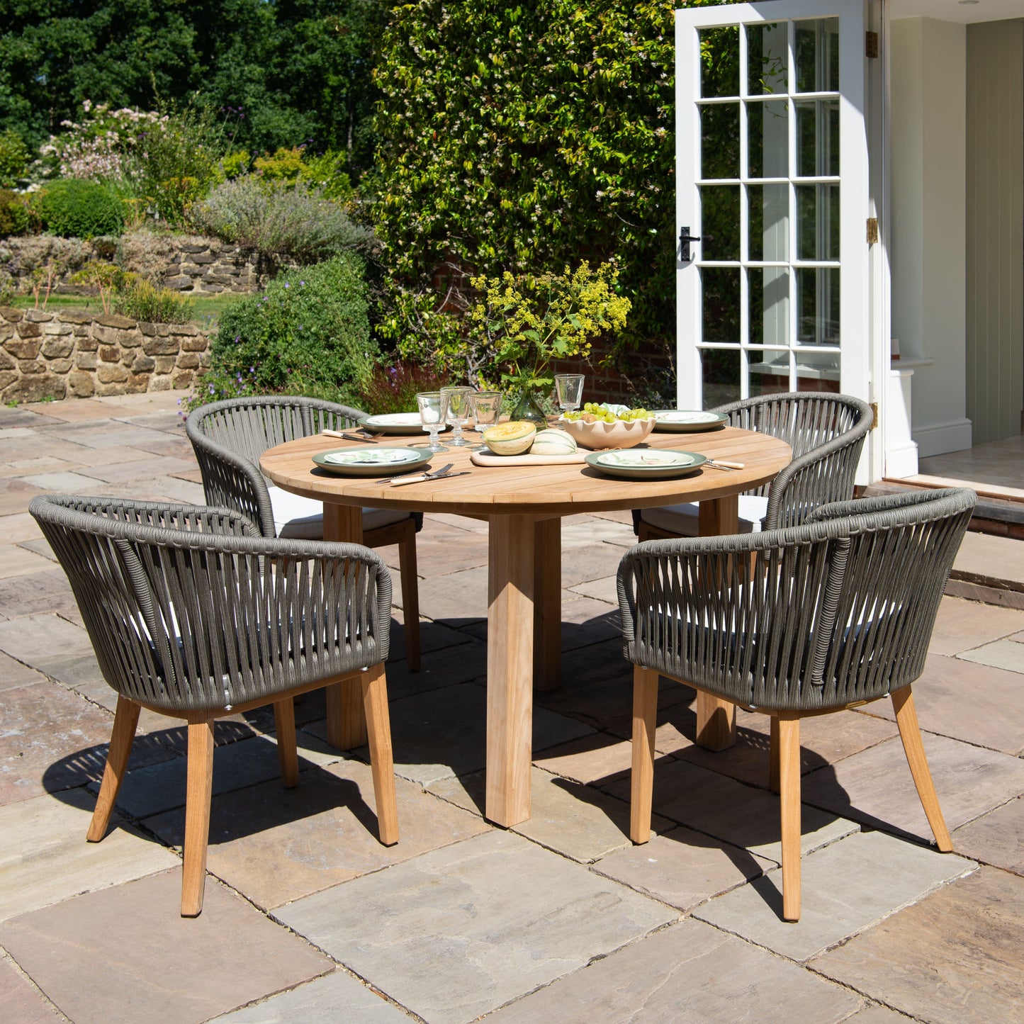 Outdoor dining setup with round teak table and four teak and rope chairs on a stone patio.
