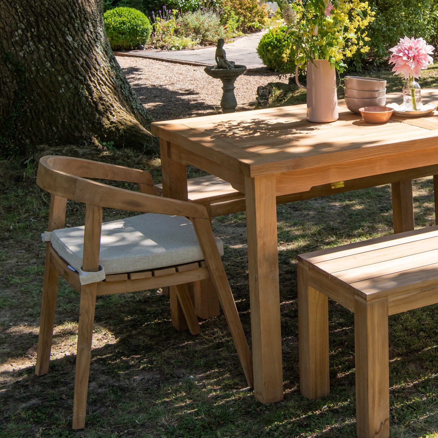 Teak wooden outdoor dining set with table, chairs, and bench under a tree in a garden.
