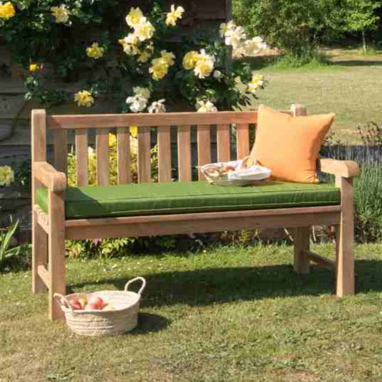 
                  
                    A classic teak garden bench with a green seat pad and an orange cushion placed on it, situated in a garden setting with a basket of fruit beside it.
                  
                