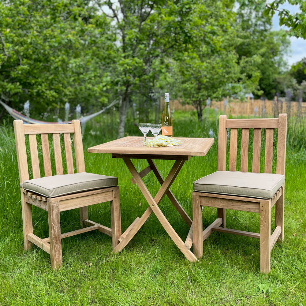 
                  
                    Two teak outdoor dining chairs with oatmeal coloured seat pads. In between is a teak square folding table with a bottle of wine and two glasses, in garden setting. 
                  
                