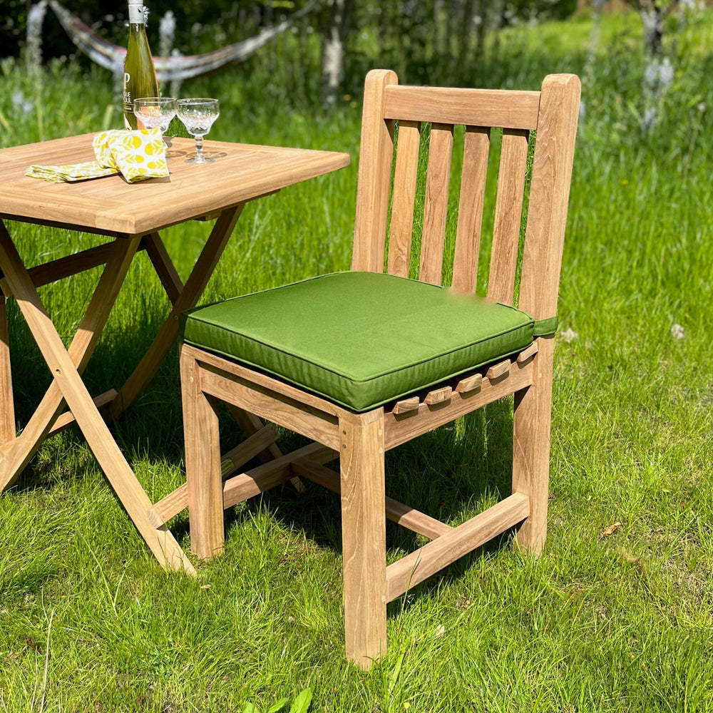 
                  
                    single teak dining chair with bright green seat cushion, next to small square folding teak table with glasses and wine, in garden setting. 
                  
                