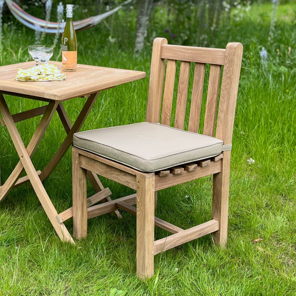 
                  
                    Single teak dining chair with grey green seat cushion, next to teak folding table with wine and glasses, in garden setting
                  
                