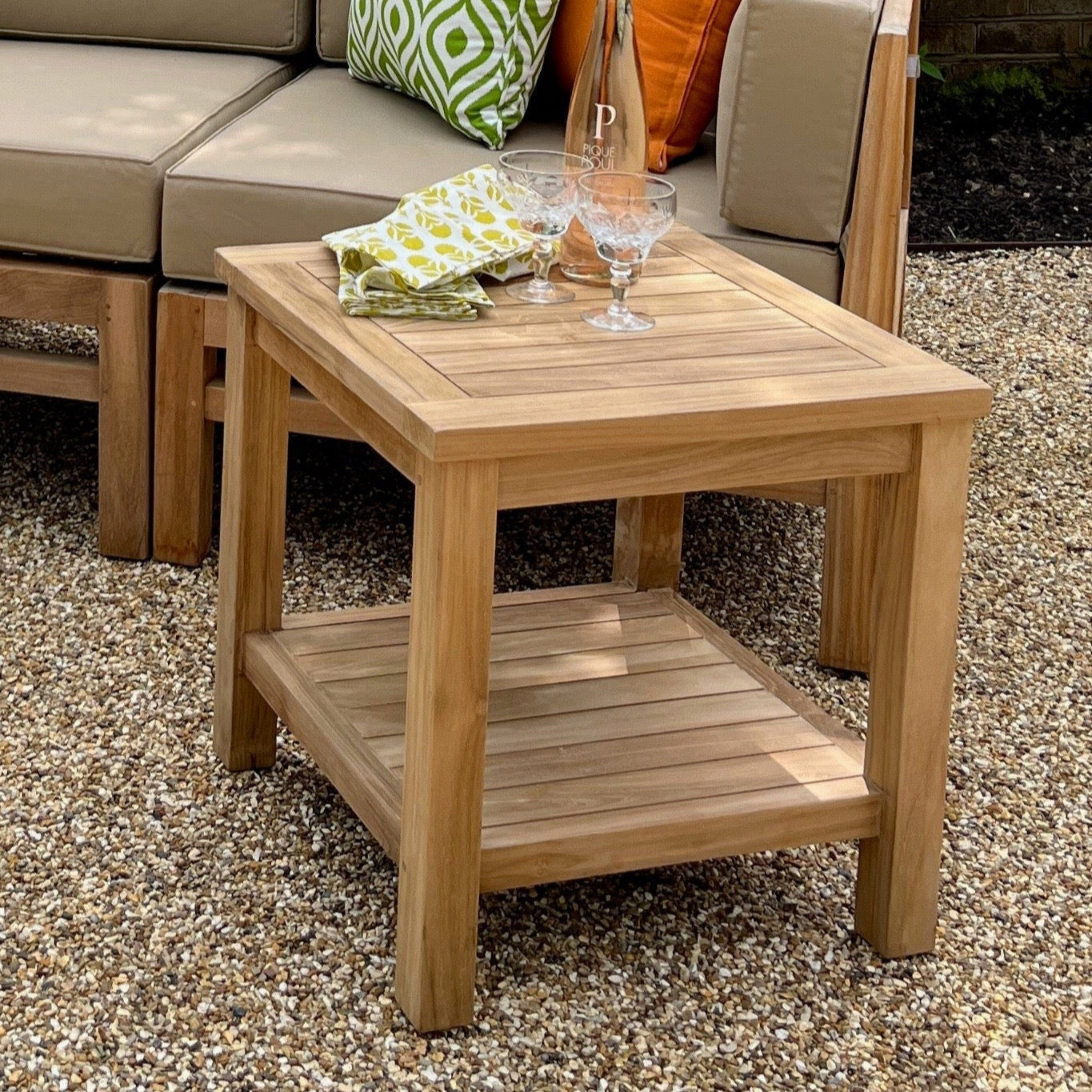 A square teak wood coffee table with a lower shelf, placed on a gravel surface with outdoor furniture in the background.