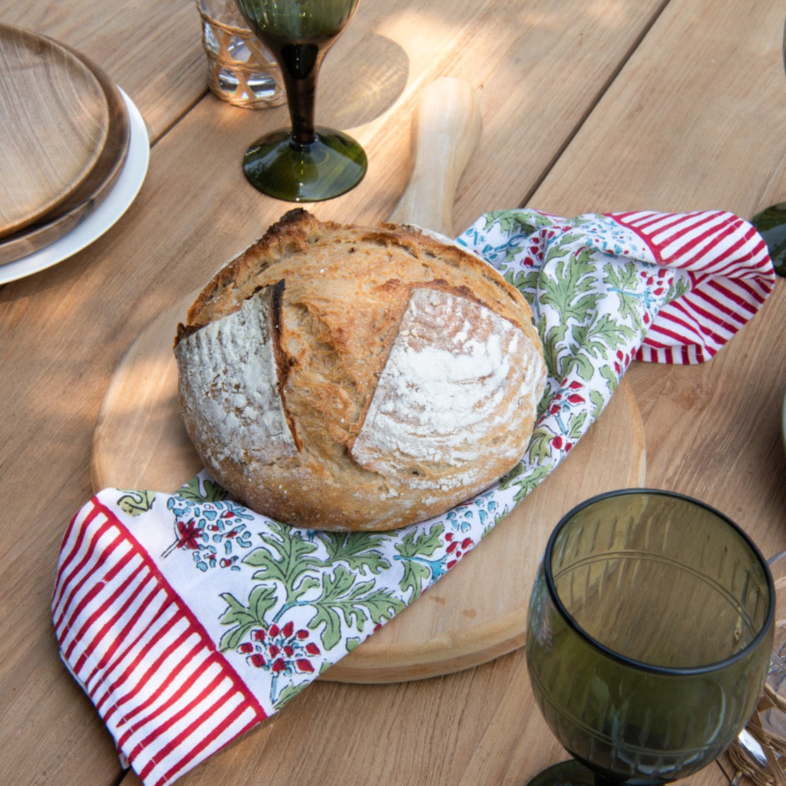 Round pizza paddle made of teak wood holding loaf of bread, on pattterned cloth, on top of teak table with plates and glasses around