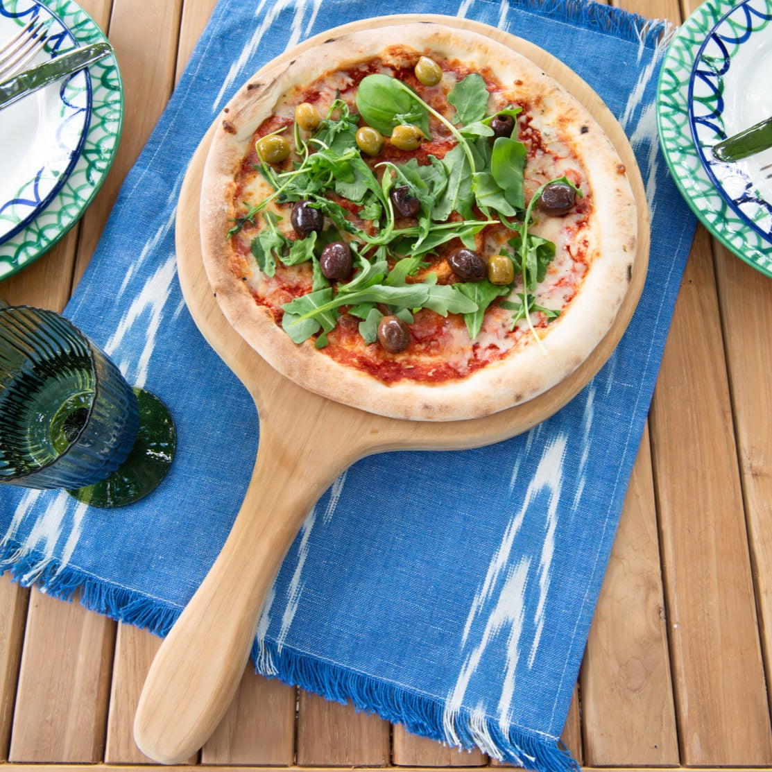 Round pizza paddle holding a pizza, sitting on blue cloth, on teak table with plates and glasses around 