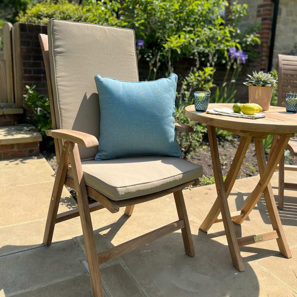 
                  
                    Single teak outdoor chair with armrests, grey seat and backrest cushion and blue scatter cushion, next to teak round folding table, on stone patio. 
                  
                