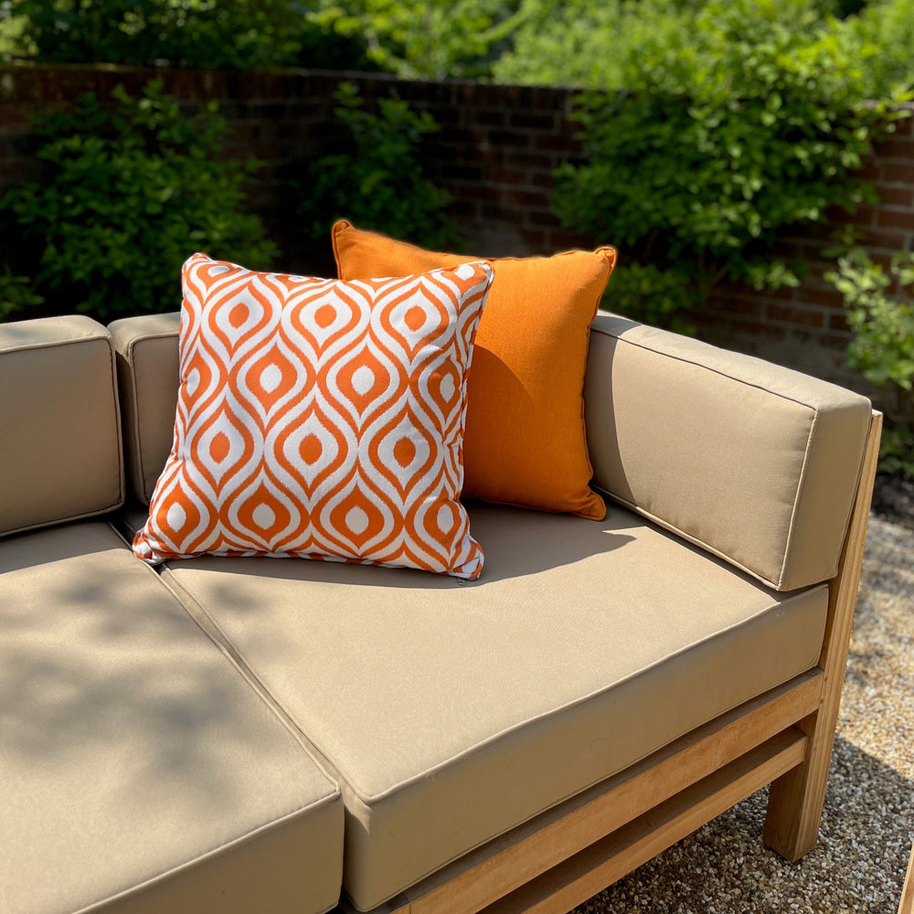 
                  
                    One end of teak outdoor sofa with oatmeal coloured seat, back and side cushions, plus plain and pattened orange scatter cushions, in garden setting.  
                  
                
