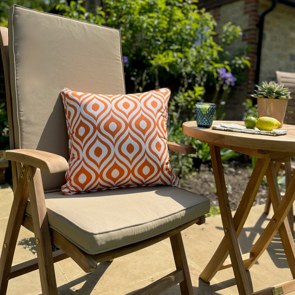 
                  
                    Single teak outdoor chair with armrests, oatmeal coloured seat and backrest cushion and orange patterned scatter cushion, next to teak round folding table, on stone patio. 
                  
                
