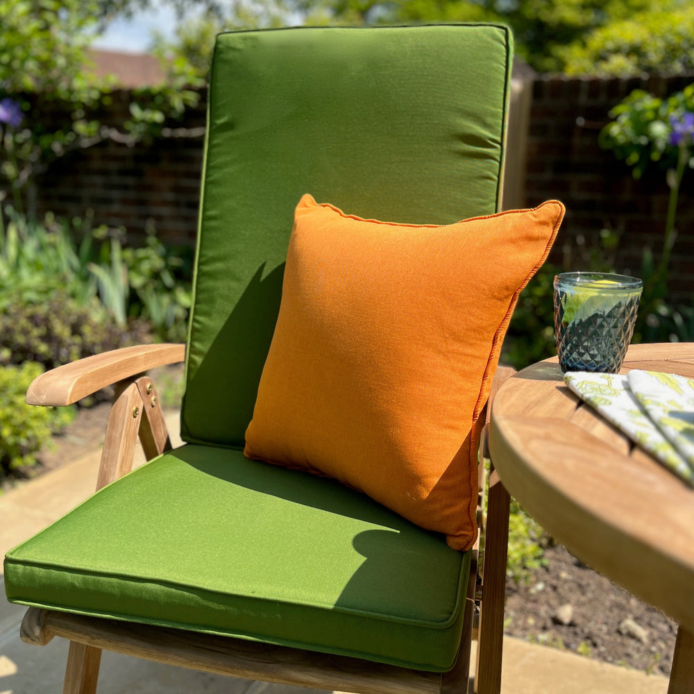 
                  
                    Teak outdoor chair with bright green seat and backrest cushions and orange scatter cushion, next to round teak table with water glass, flowerbeds and brick wall in background.   
                  
                