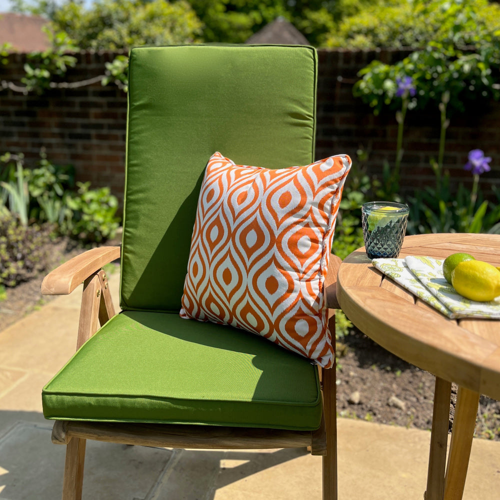
                  
                    Teak outdoor chair with bright green seat and backrest cushions and orange scatter cushion, next to round teak table with water glass and fruit, with brick wall in background.   
                  
                