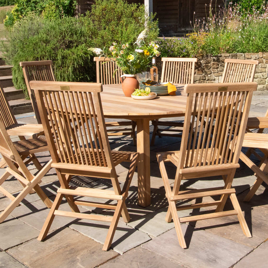 Teak wooden outdoor dining set on a stone patio with a garden and gazebo in the background.