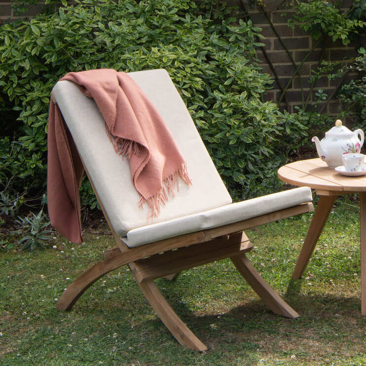Two teak wooden reclining chairs with white cushions and a small round teak table in a garden setting.
