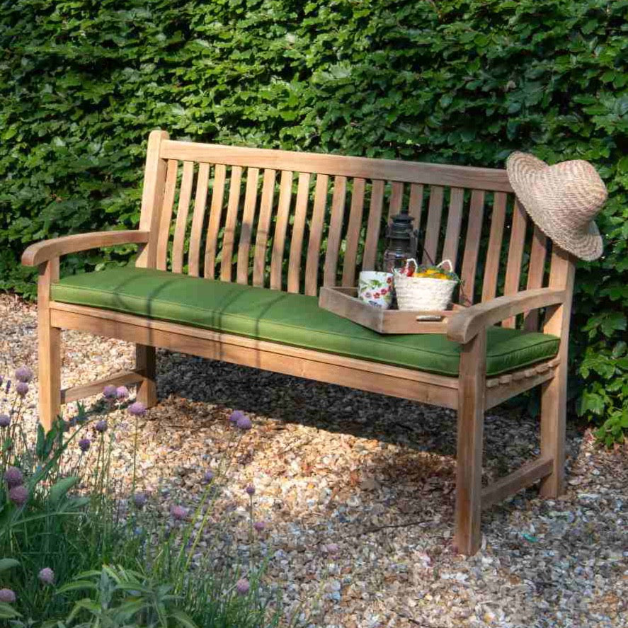 
                  
                    A teak garden bench with green cushions placed on a gravel path, featuring a hat and a teak serving tray with a cup and basket, on top of the cushion. 
                  
                