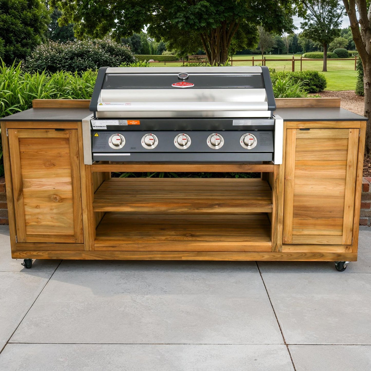 Outdoor barbecue with teak cabinets either side and shelves beneath, on a patio with garden in the background