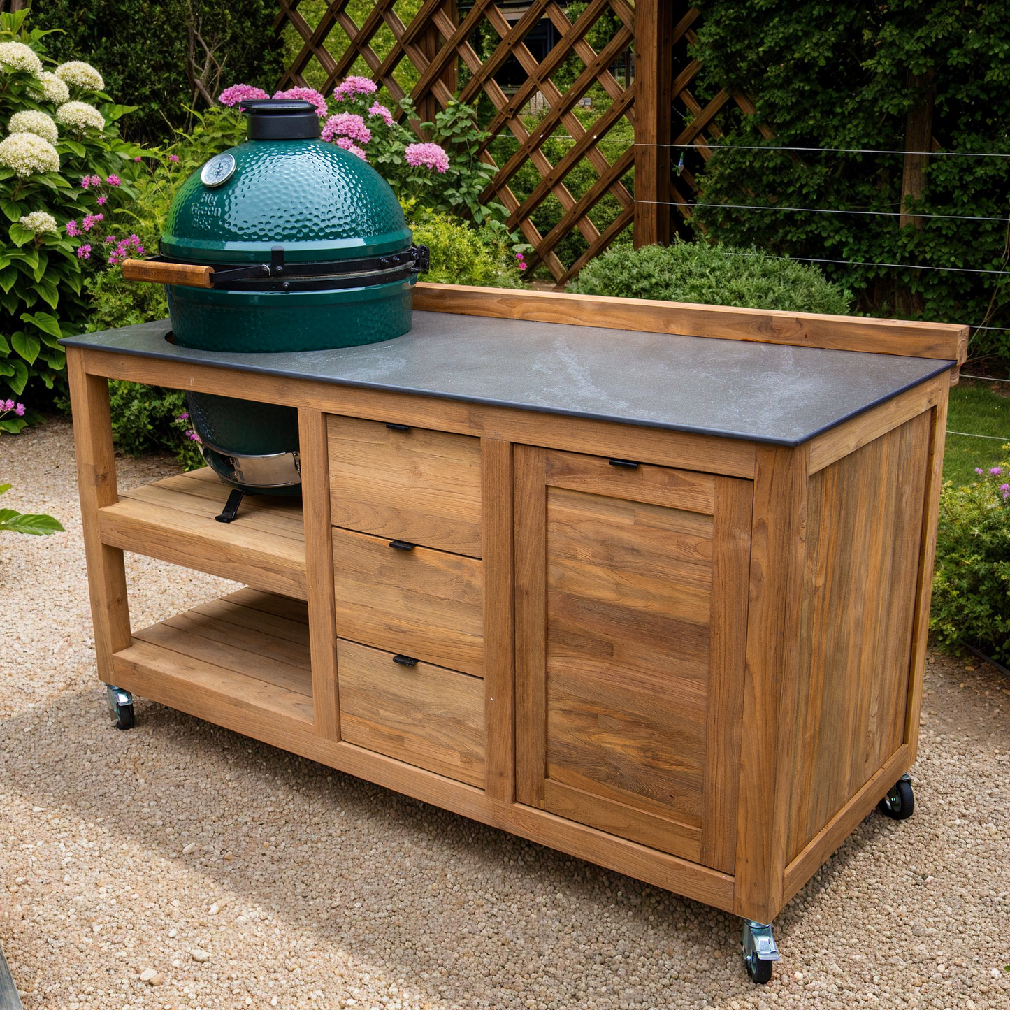 Teak outdoor kitchen island with a green barbecue grill on a gravel patio surrounded by plants.