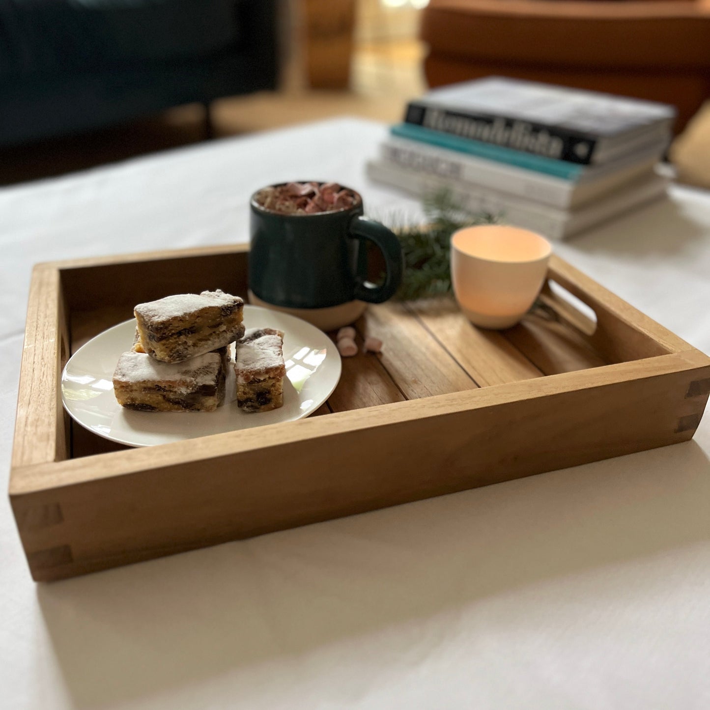 Teak wood serving tray with high sides, set with plate of cakes and coffee cup, on white tablecloth, with stack of books in the background