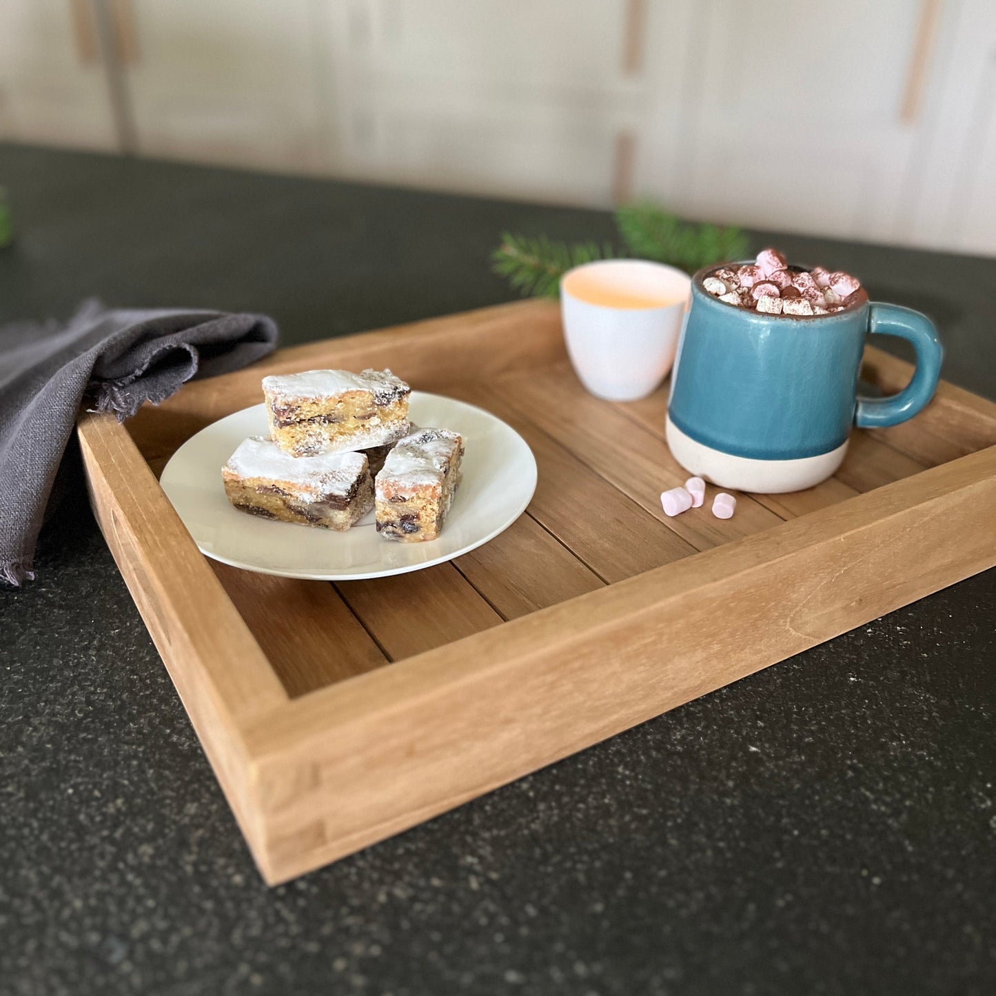 A rectangular teak serving tray with handles, displaying a plate of food and a cup on top, placed on a dark surface with a sprig of greenery in the background.