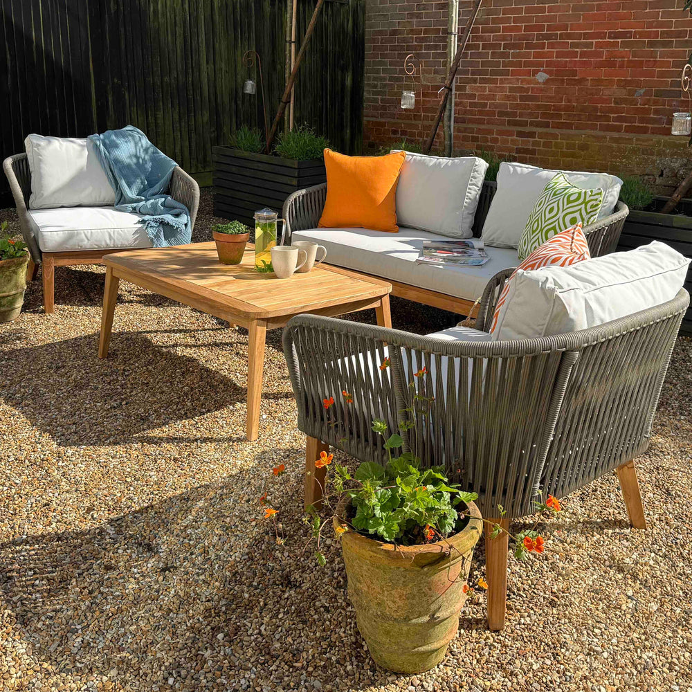 
                  
                    Two seater outdoor sofa and two armchairs, all made from teak and rope with white seat cushions, on gravel patio with brick wall behind. 
                  
                