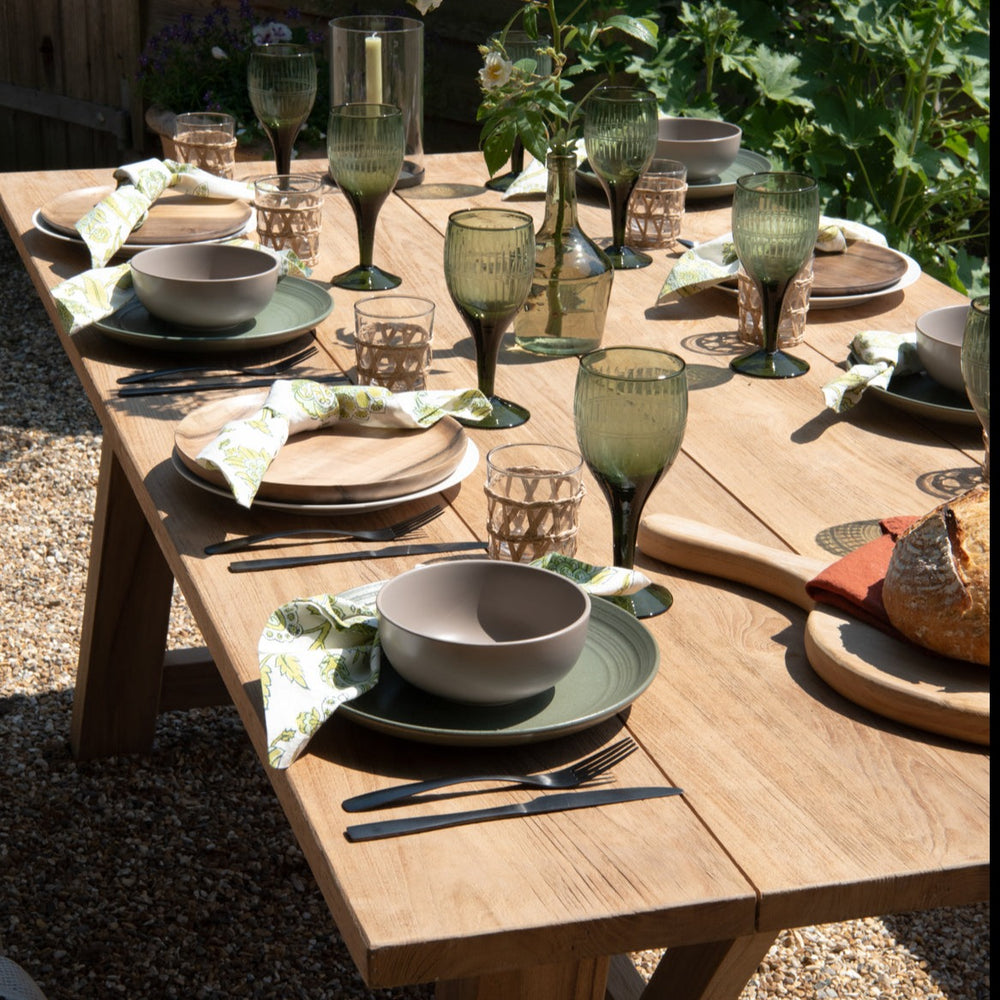 Teak rectangular table with six place settings and wine glasses, on gravel patio with shrubs in the background