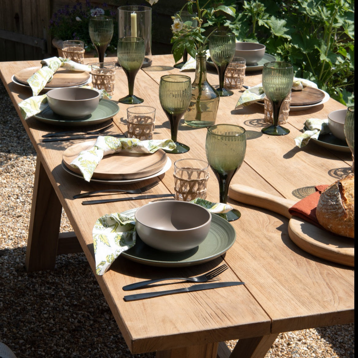 Teak rectangular table with six place settings and wine glasses, on gravel patio with shrubs in the background