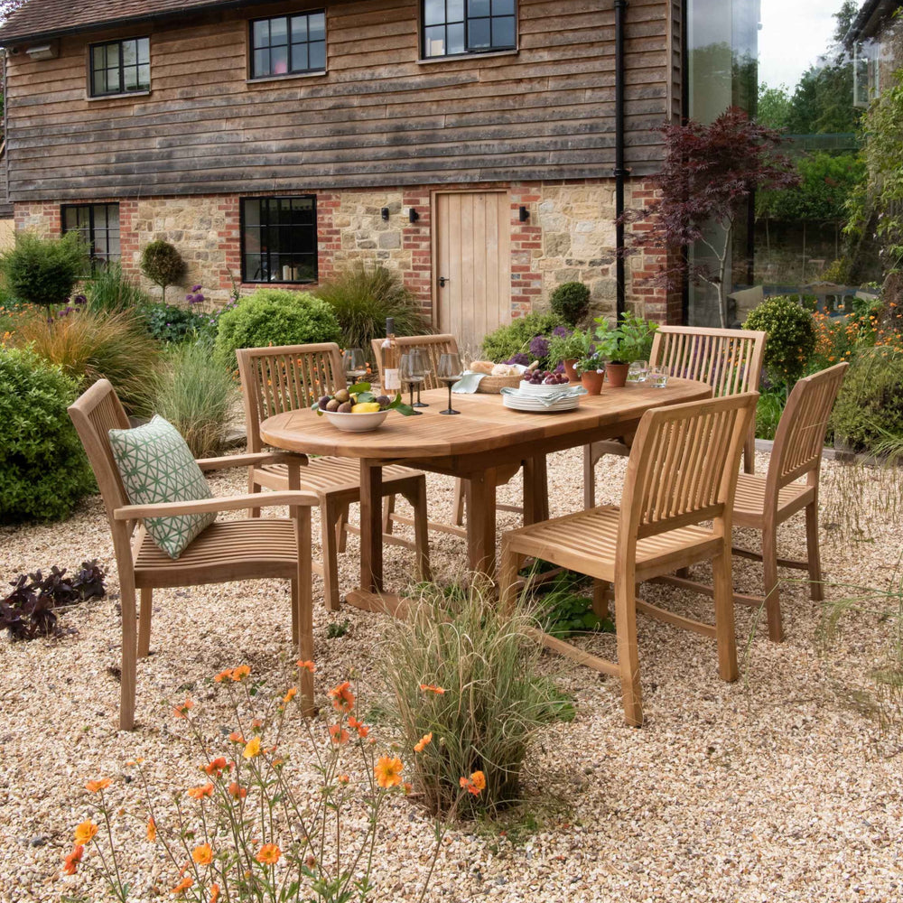 
                  
                    Teak oval dining table with plants, glasses and fruit, with teak dining chairs set around, on gravel patio with wooden, redbrick building behind.  
                  
                