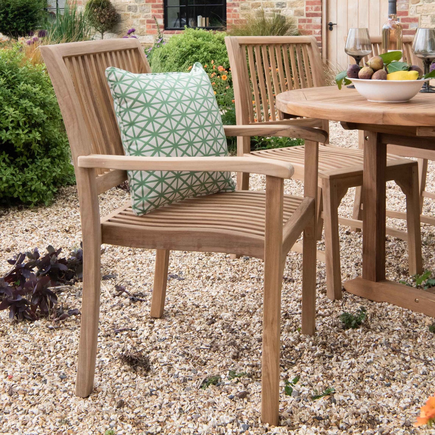 Teak outdoor dining chairs with armrests and green patterned scatter cushion, next to teak, oval dining table with fruit and wine glasses, on gravel patio. 