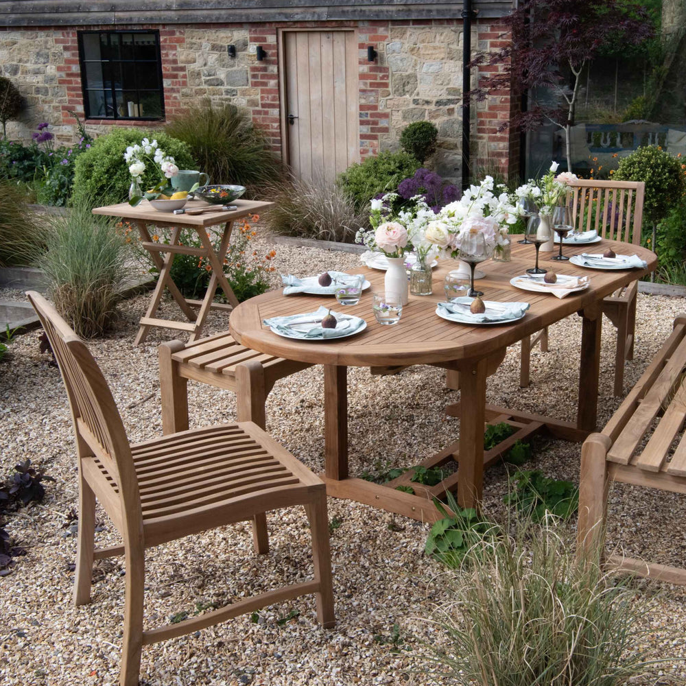 
                  
                    Teak oval dining table, set with eight place settings and flowers, with teak benches and dining chairs, square teak folding table to the side, on gravel patio with stone building behind. 
                  
                