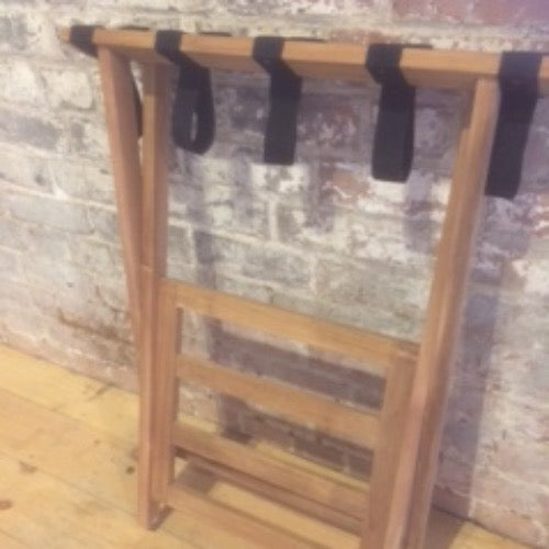 Teak luggage rack with teak wood frame, shelf beneath and canvas straps to hold bags, folded flat and leaning against brick wall. 