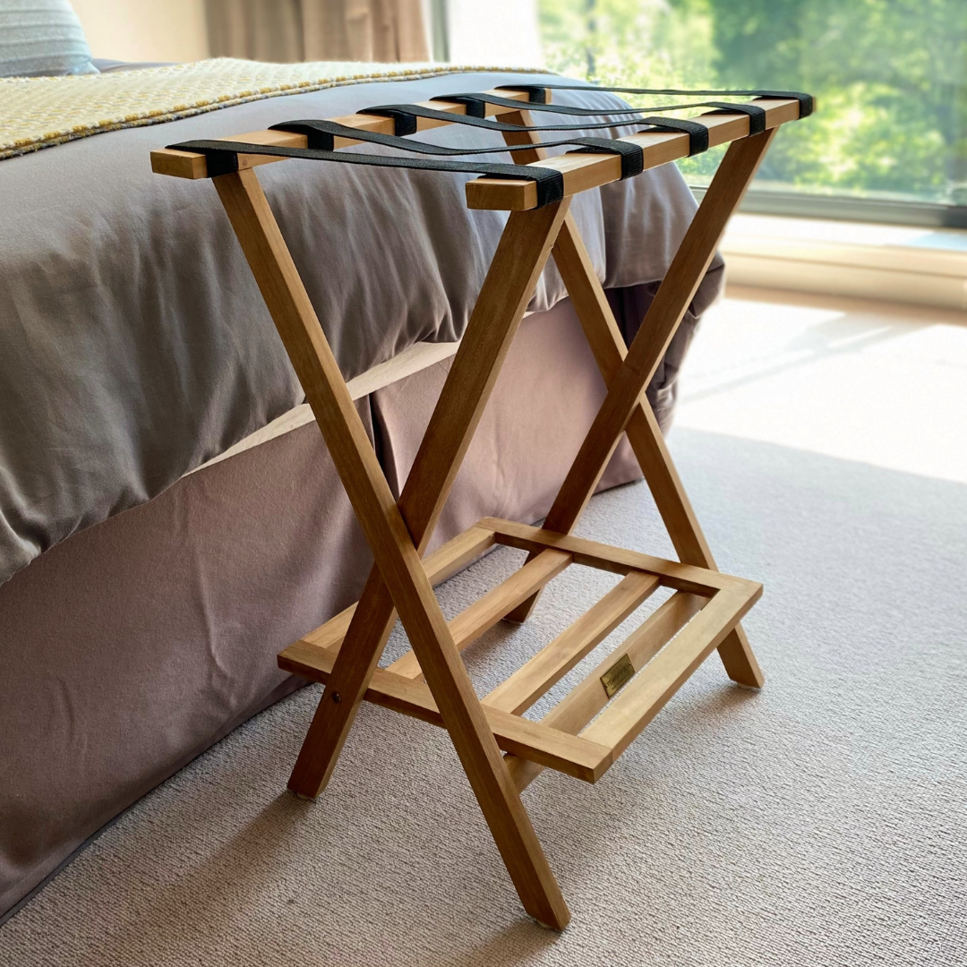 teak folding luggage rack with canvas straps to hold luggage and shelf below, at foot of bed on carpet with window in the background. 