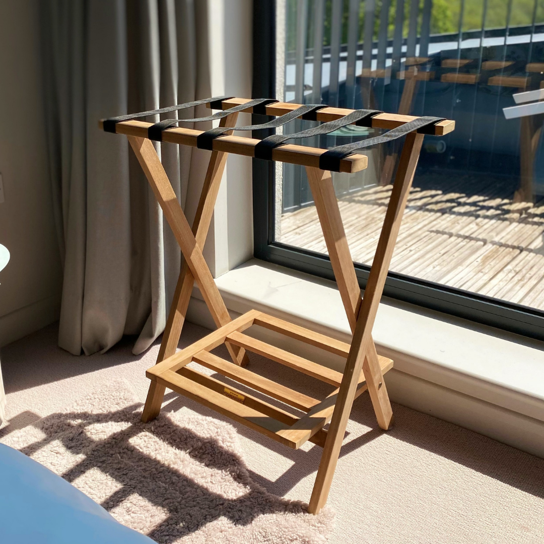Teak folding luggage rack with canvas bands on top to hold luggage, and teak slatted shelf beneath, in front of window, on carpeted floor. 