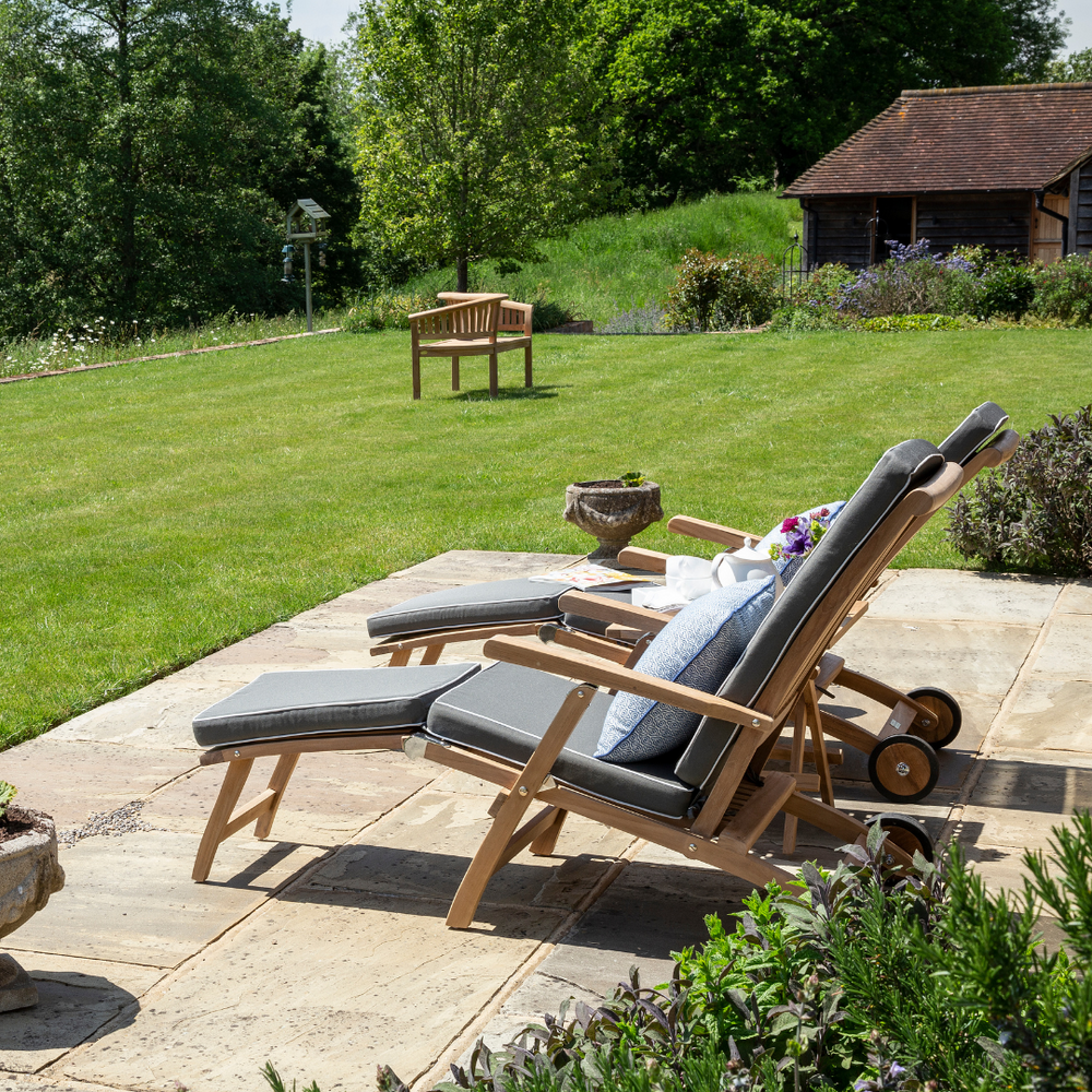 
                  
                    Two teak reclining steamer chairs with armrests and green cushions, on patio facing lawn, with teak love seat and garden building in background 
                  
                