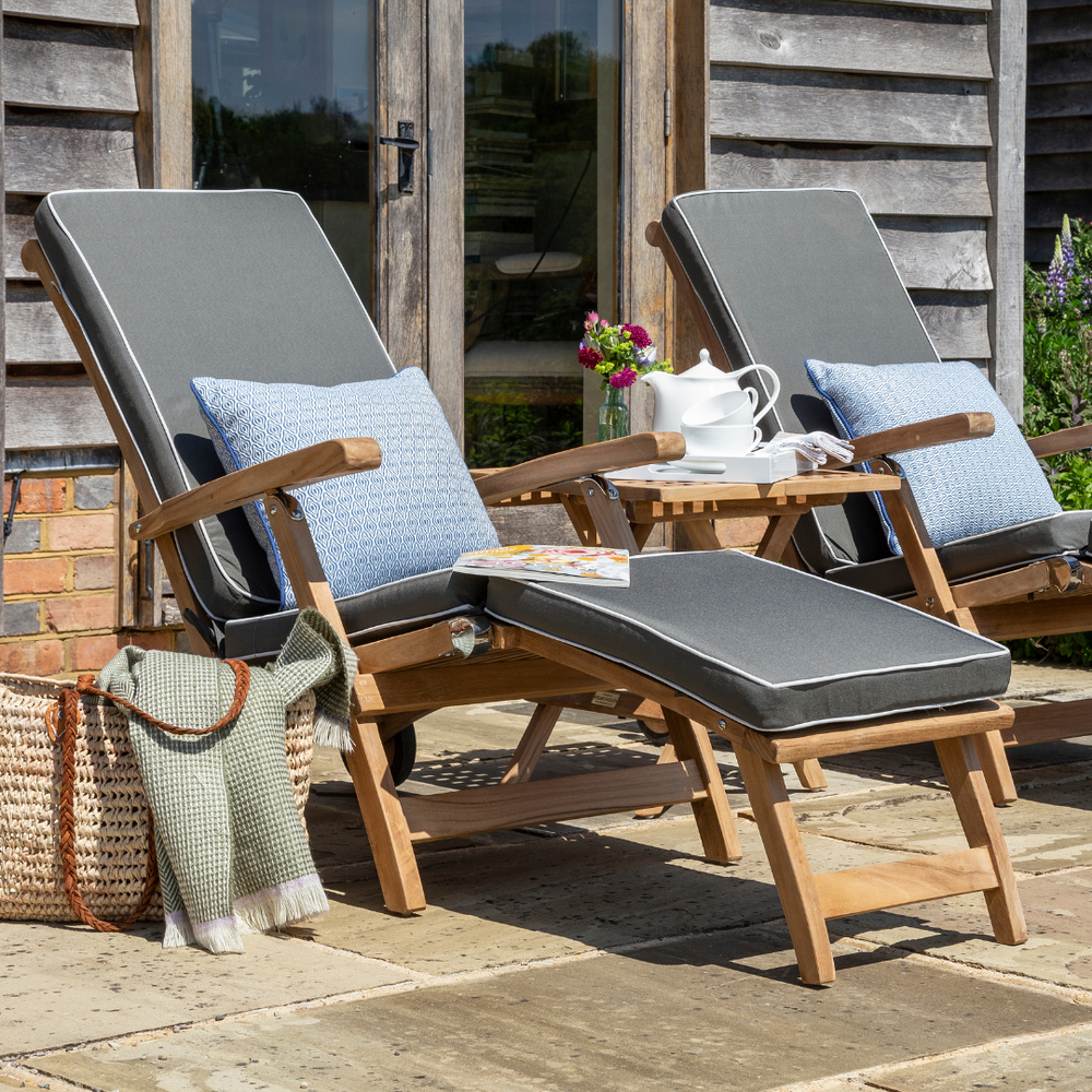 
                  
                    Two reclining teak wood steamer chairs with built-in footstools and armrests, equipped with wheels, on a patio, with cushions in a matching colour
                  
                
