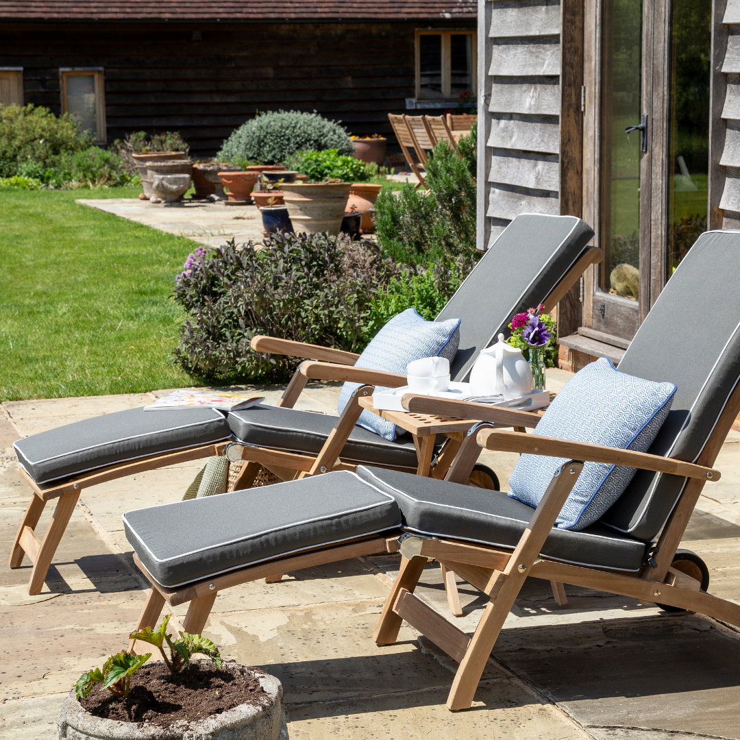 Two reclining teak wood steamer chairs with built-in footstools and armrests, equipped with wheels, on a patio, with cushions in a matching colour