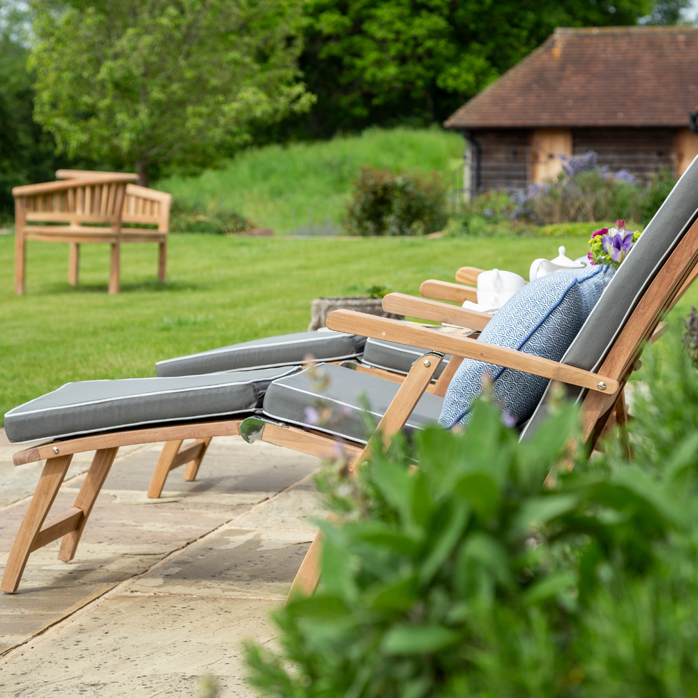 
                  
                    Two teak reclining chairs with built in armrests and footstools, green full length cushions, in garden setting with teak love seat in background. 
                  
                