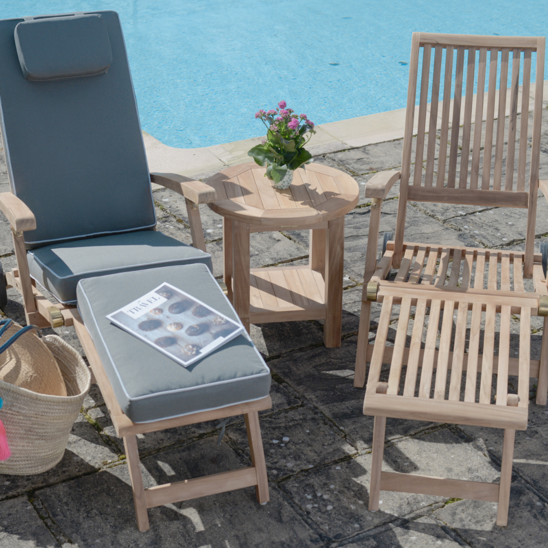Teak reclining steamer chairs, on stone patio, in front of swimming pool. One chair with grey, full-length cushion, one without. A small, round teak coffee table sits between. 