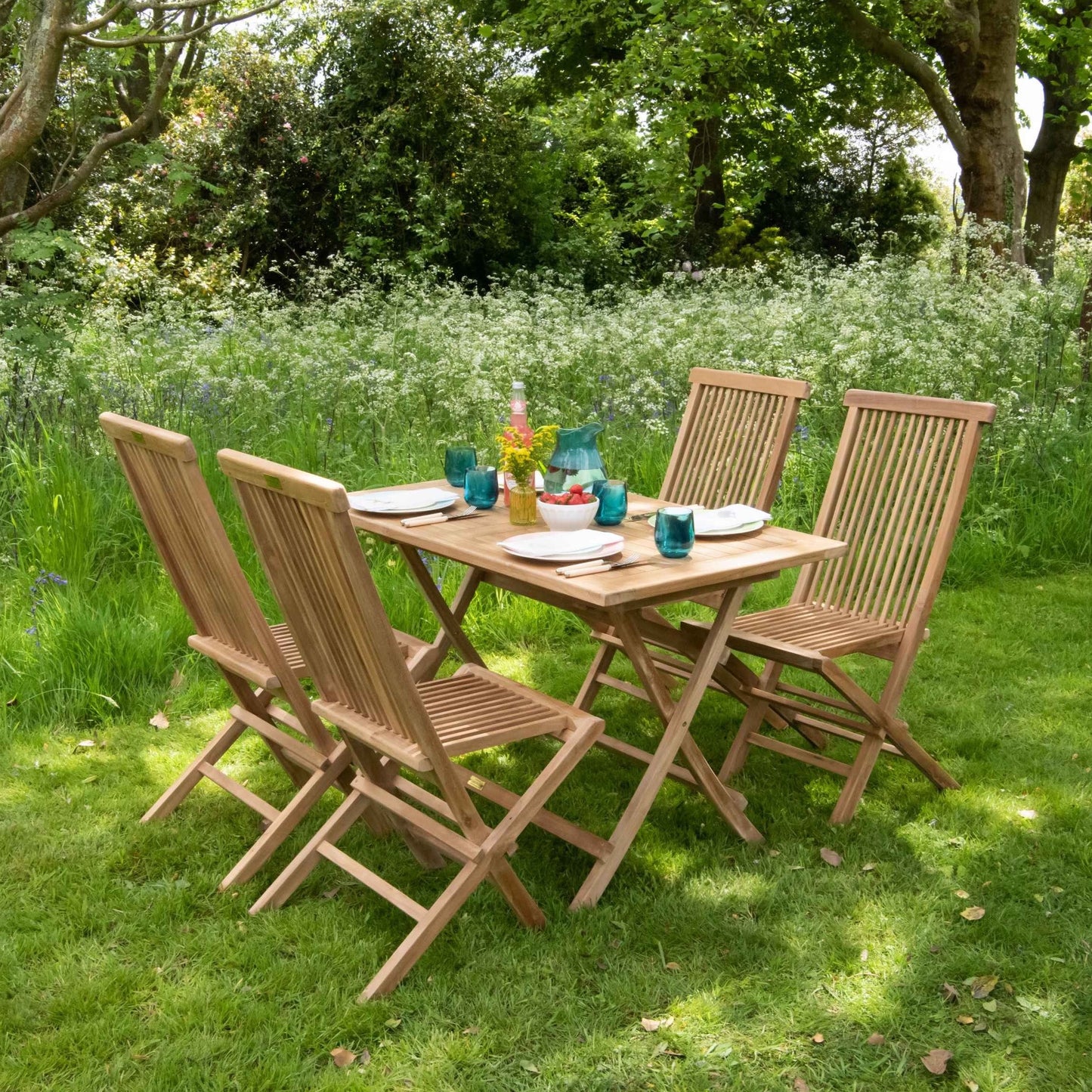 Teak folding rectangular table, laid with green glassware and plates, with teak folding chairs around, in garden setting.  