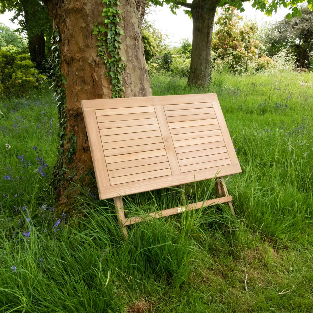 
                  
                    Teak folding rectangular table, in folded position, leaning against tree trunk, in garden setting with trees behind. 
                  
                