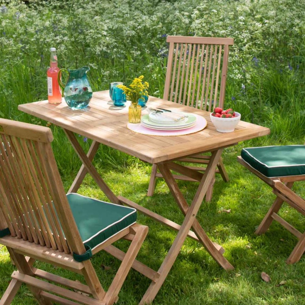 
                  
                    Teak Folding Rectangular Table with teak folding chairs set around in a garden setting. The chairs have green and white seat cushions. The table is holding fruit, plates and glasses. 
                  
                