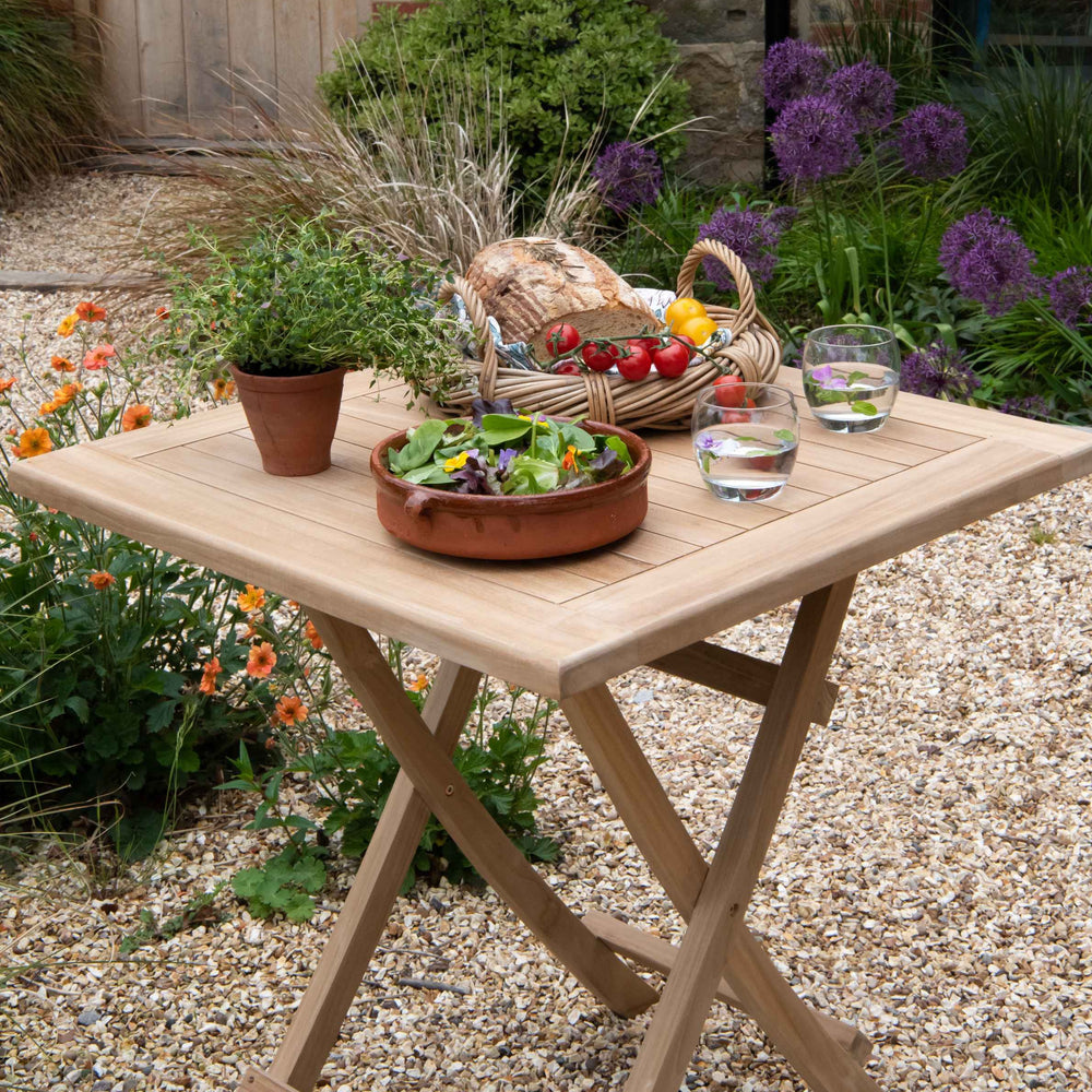 Teak square folding table with basket of bread and tomatoes, salad bowl and water glasses, on gravel with flowerbeds behind. 
