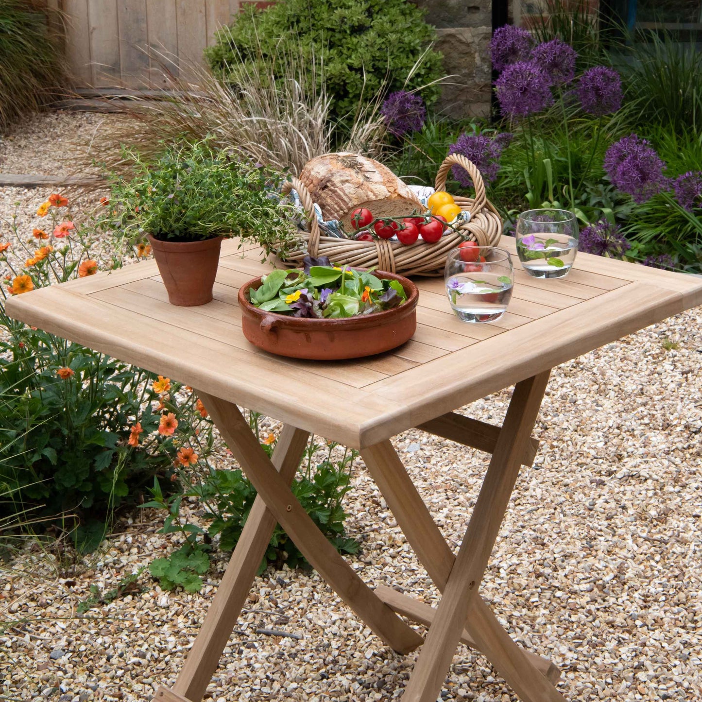 Teak square folding table with basket of bread and tomatoes, salad bowl and water glasses, on gravel with flowerbeds behind. 