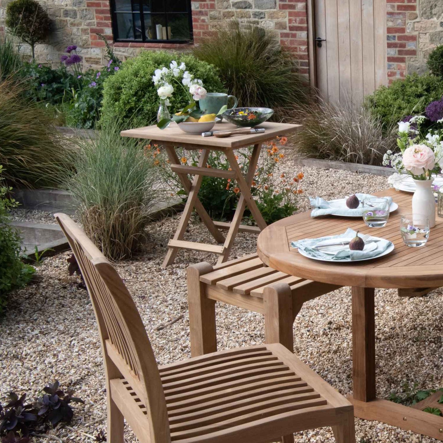 Teak Square Folding Table holding fruit and flowers with oval teak dining table and chairs in foreground, on a gravel surface with shrubs around.    