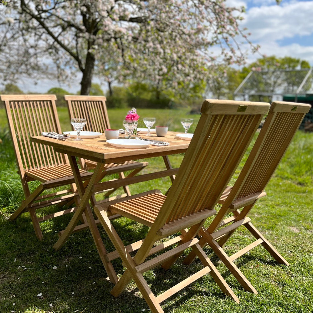 
                  
                    Teak Folding Rectangular Dining Table laid with place settings, with four teak folding chairs, in a garden setting. 
                  
                