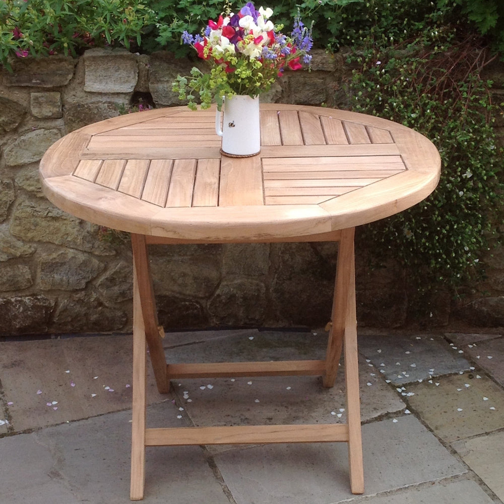 
                  
                    Round folding teak table holding white jug of flowers, on a stone patio with brick wall behind.  
                  
                